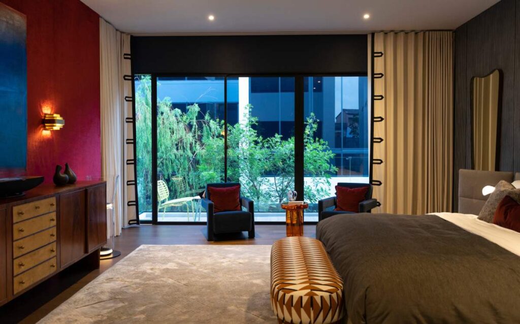 Interior de dormitorio con un diseño moderno y retro de mediados de siglo. Aparece una cama matrimonial, dos butacas y una cómoda de madera en la pared.