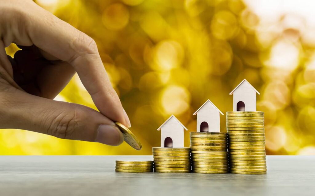 Mano apilando monedas junto a pequeñas casas de juguete, simbolizando el ahorro para la vivienda.
