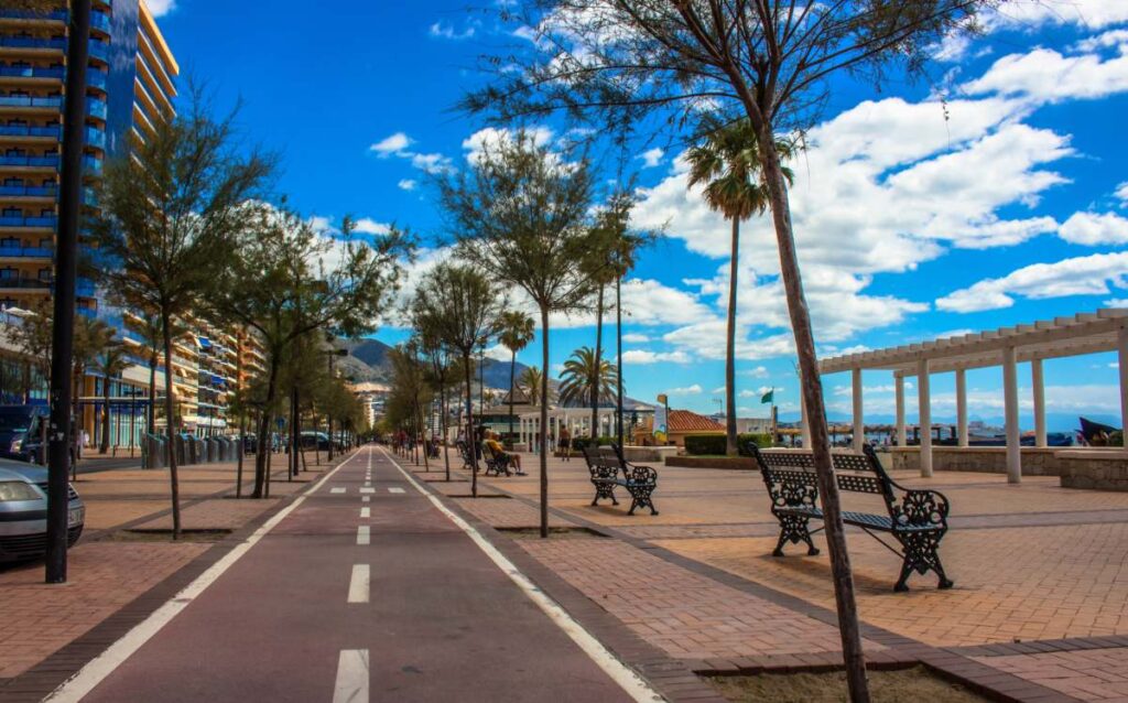 Paseo marítimo de Fuengirola con carril bici rojo, palmeras, bancos de forja y edificios de apartamentos bajo un cielo azul con nubes