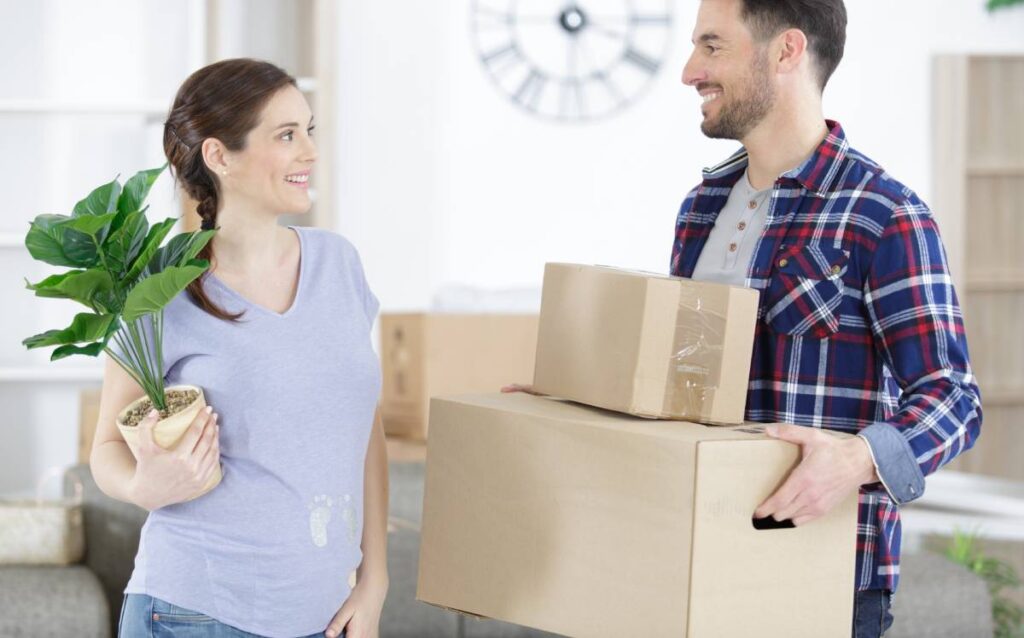 Pareja sonriente cargando cajas de cartón y una planta durante una mudanza en su nuevo hogar