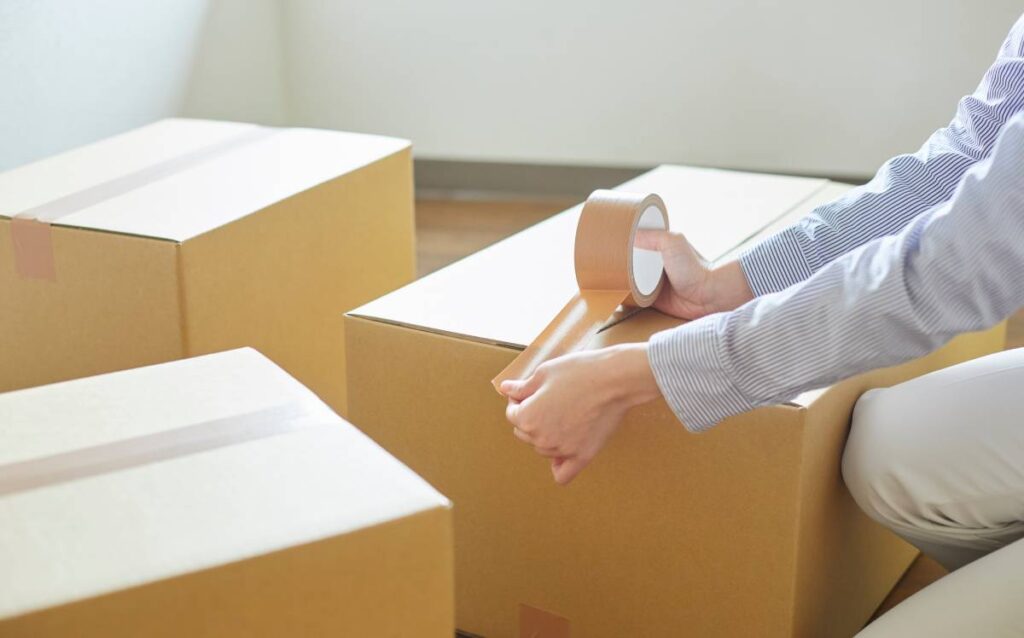 Manos de una persona usando cinta adhesiva marrón para cerrar una caja de cartón de mudanza, con otras cajas al fondo
