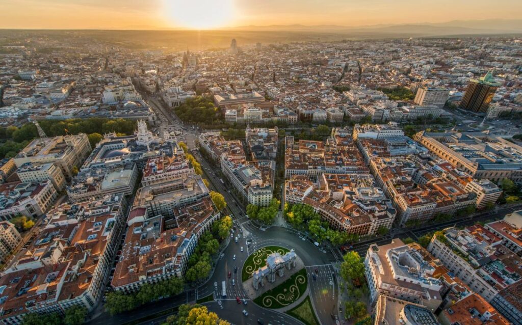 Vista aérea panorámica de Madrid al atardecer, destacando la Puerta de Alcalá en primer plano y la Gran Vía extendiéndose hacia el horizonte