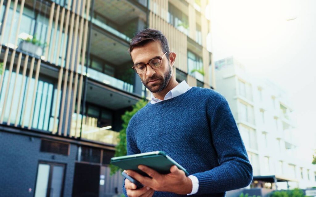Hombre con gafas y jersey azul utilizando una tableta frente a un edificio de oficinas moderno