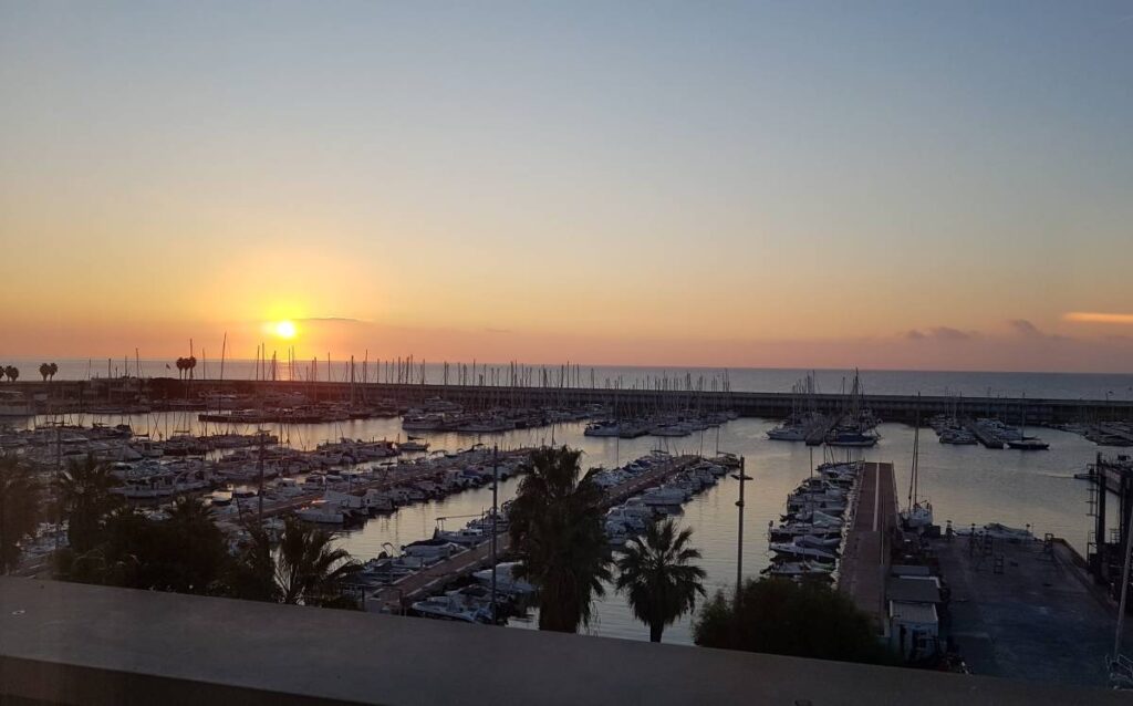 Vista aérea elevada de un gran puerto deportivo lleno de yates y veleros al atardecer, con el sol poniente en el horizonte