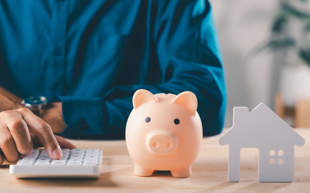 Persona utilizando una calculadora junto a una hucha de cerdito y una casa de juguete