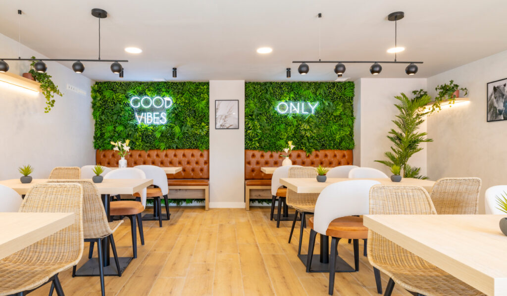 Imagen de un acogedor local que destaca por su diseño moderno y luminoso que incluye paredes verticales de plantas con carteles de neón, mobiliario de madera, lámparas esféricas y asientos tapizados.