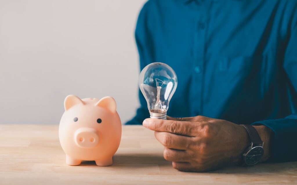 Imagen sobre el ahorro energético, una mano de un hombre sosteniendo un bombillo convencional apagado junto a una hucha con forma de cerdito. 