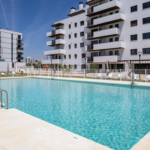 Piscina comunitaria de Célere Arce rodeada de zonas ajardinadas, solárium con tumbonas y modernos edificios residenciales con amplias terrazas y espacios comunes.