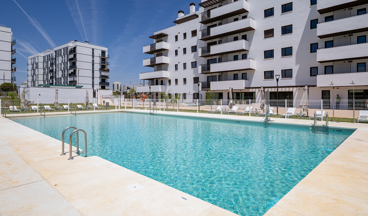 Piscina comunitaria de Célere Arce rodeada de zonas ajardinadas, solárium con tumbonas y modernos edificios residenciales con amplias terrazas y espacios comunes.
