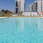 Área de piscina comunitaria de Célere Arce integrada en un entorno residencial moderno, con zonas verdes, espacios de ocio exterior y edificios de diseño contemporáneo al fondo.