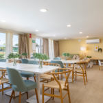 Sala comunitaria de Célere Arce con mesas, sillas, zona de descanso, espacio infantil y amplios ventanales en un entorno moderno y multifuncional.