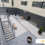 La imagen muestra una zona común interior de Célere Arts II en julio de 2026, con escaleras metálicas de acceso, pavimento instalado, jardineras decorativas y áreas de tránsito peatonal prácticamente finalizadas dentro de la promoción residencial.