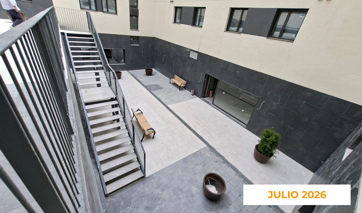 La imagen muestra una zona común interior de Célere Arts II en julio de 2026, con escaleras metálicas de acceso, pavimento instalado, jardineras decorativas y áreas de tránsito peatonal prácticamente finalizadas dentro de la promoción residencial.
