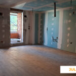 La imagen muestra una vivienda en construcción de la promoción Célere Parqueluz III en julio de 2026, con el solado ya instalado, tabiques de yeso laminado en fase de acabado, instalaciones vistas y acceso a una terraza a través de grandes ventanales.