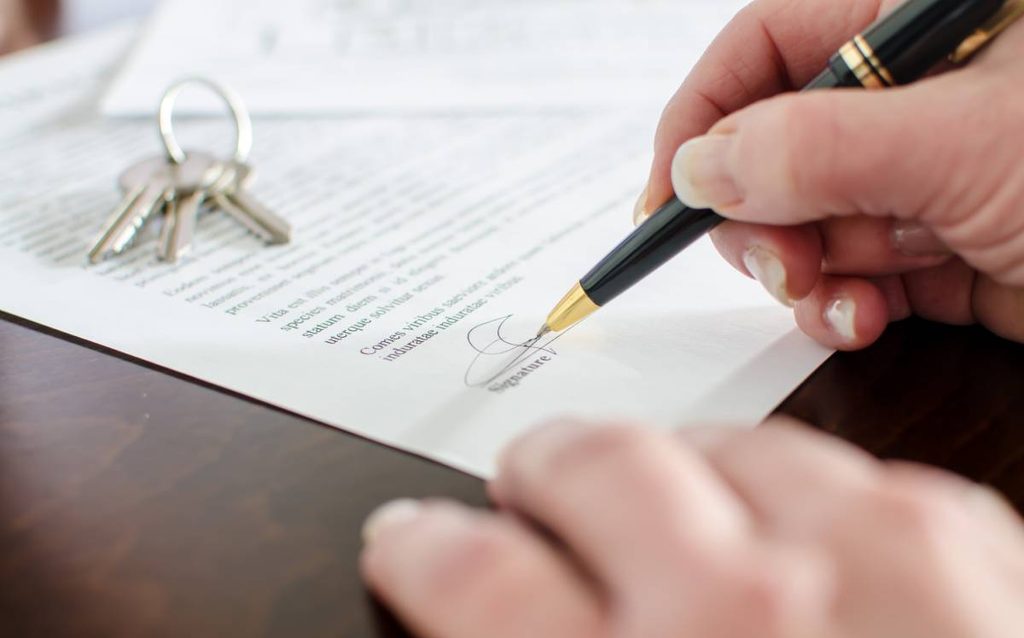 Persona firmando un contrato hipotecario junto a las llaves de una vivienda durante el proceso de compraventa