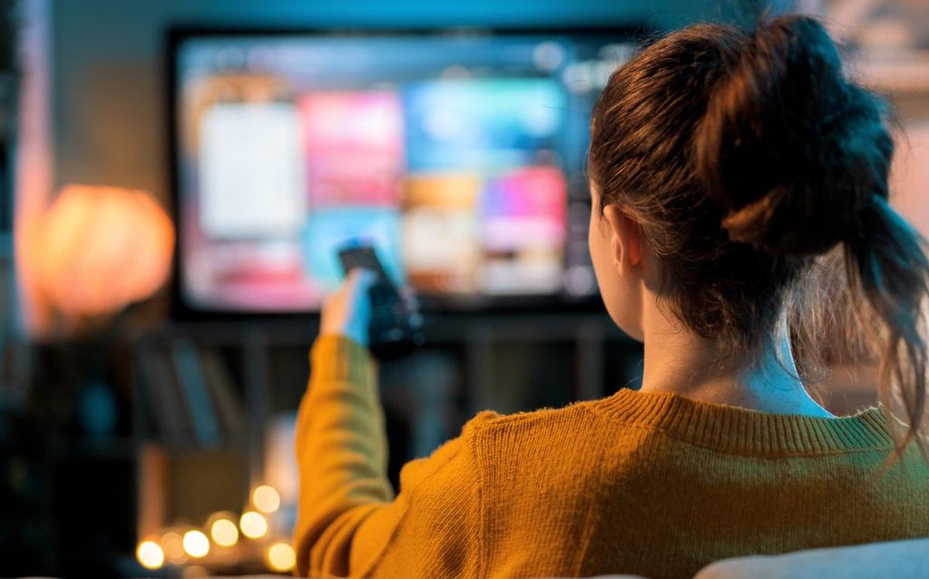 Mujer utilizando el mando a distancia frente al televisor con plataformas de streaming, uno de los gastos vampiro en el hogar
